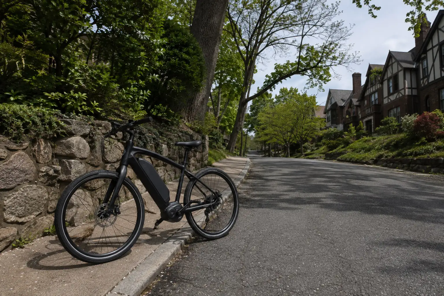 Electric-assist bicycle beside a sloping street, stone wall and Tudor homes, illustrating Riverdale NY.