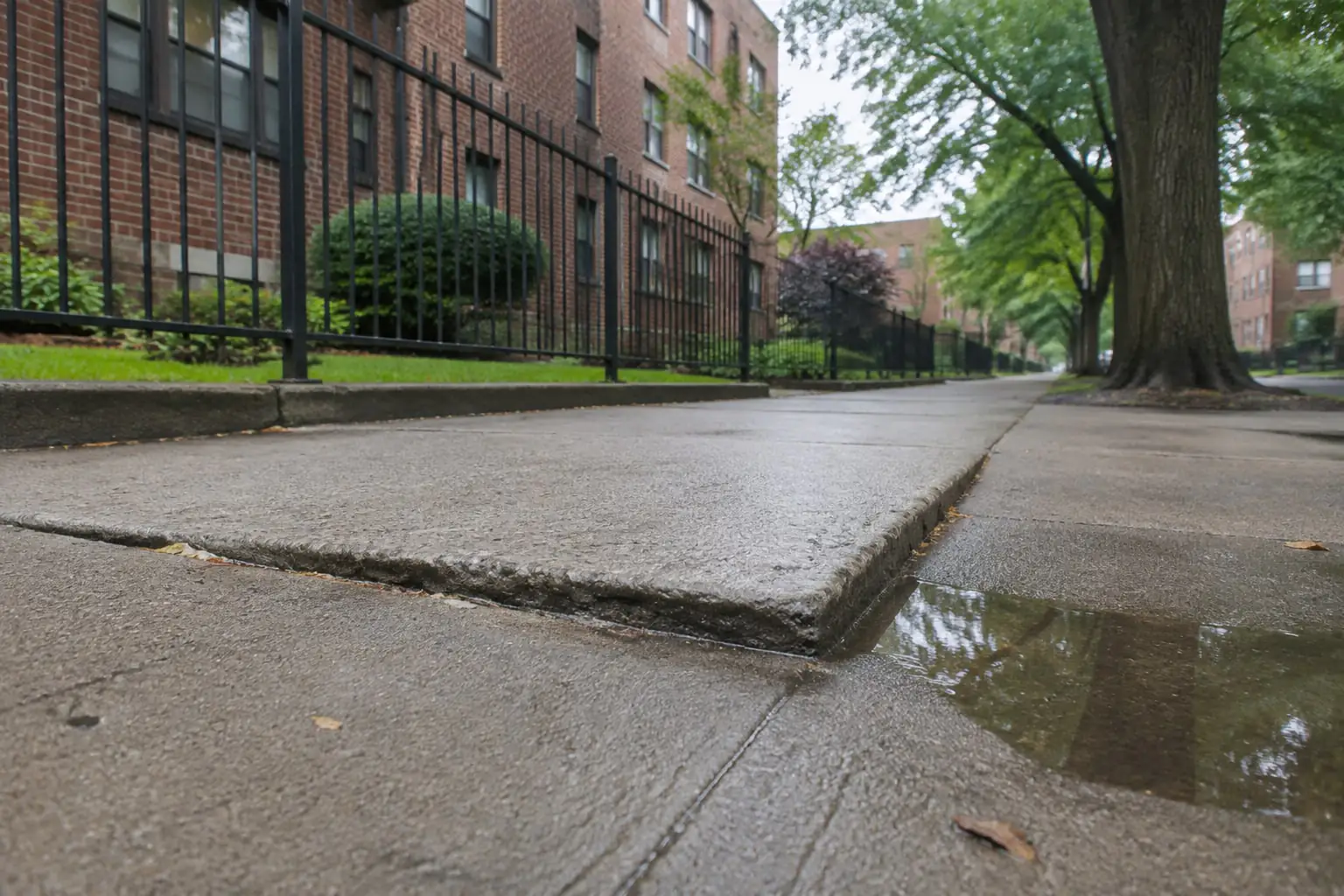 Uneven concrete sidewalk and a rain puddle beside brick apartments, illustrating a Soundview NY fall hazard.